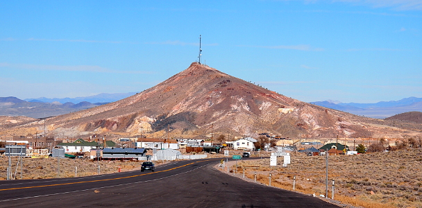 [There is a large rock-dirt hill with a tower atop it. In front of the hill are numerous single-story buildings. The image was taken from a pull-off beside the road and the asphalt of the pull-off as well as the road to town are visible. In the far distance are many mountains.]